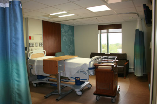 Interior patient bedroom showing hospital bed, medical equipment, window with natural light, and treatment furniture