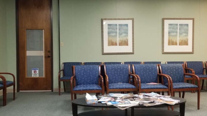 Interior waiting room with blue upholstered wooden chairs, framed landscape artwork, and door with restroom signage