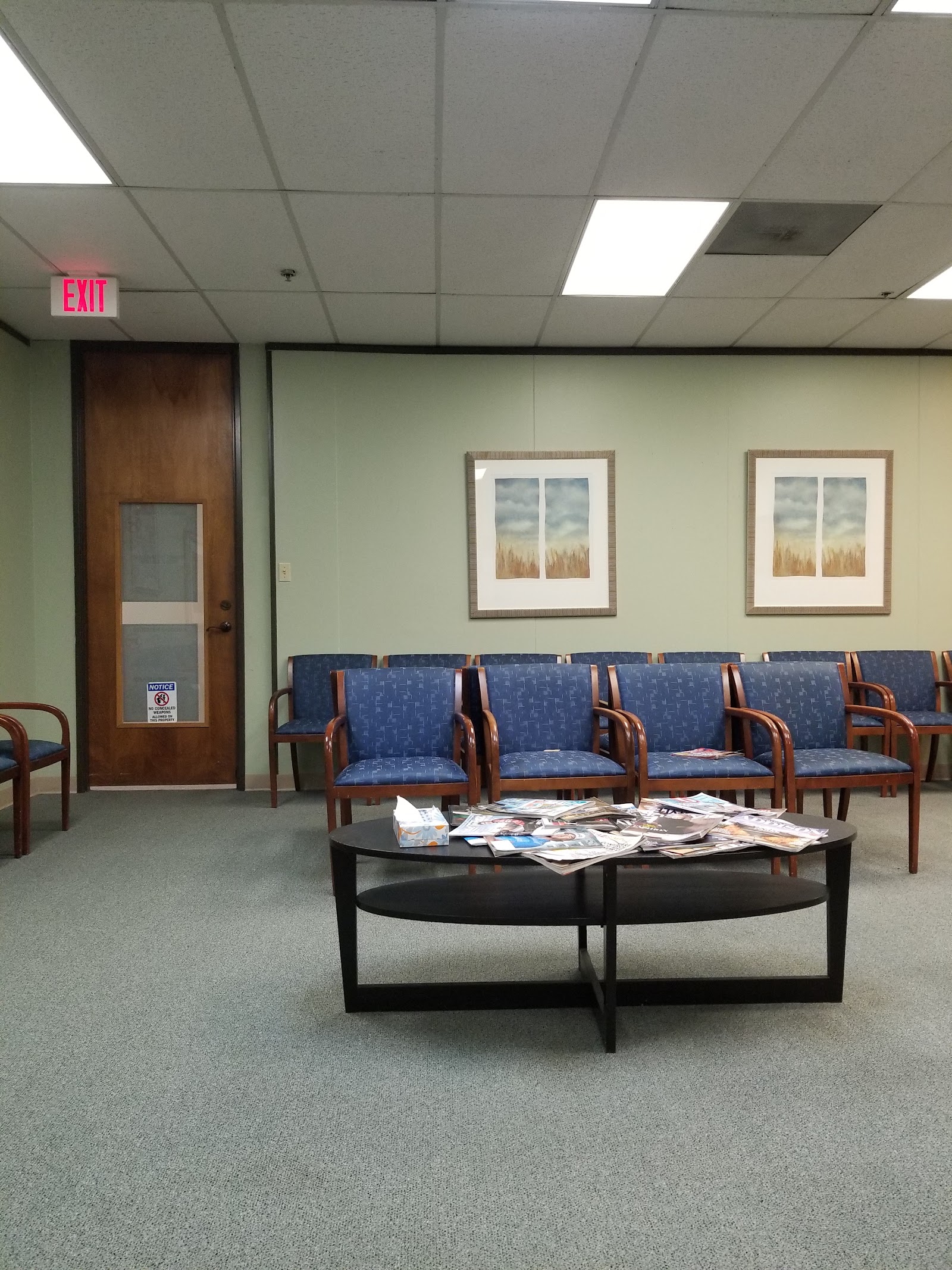 Interior waiting room with blue upholstered wooden chairs, framed landscape artwork, and door with restroom signage