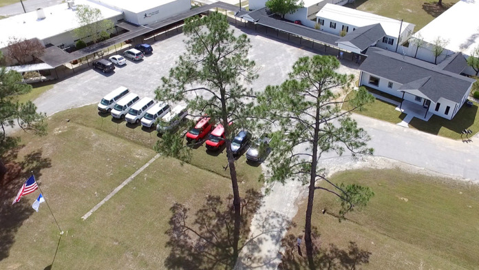 Exterior covered walkway and patio with metal framing, flowering plants, lawn, and facility buildings
