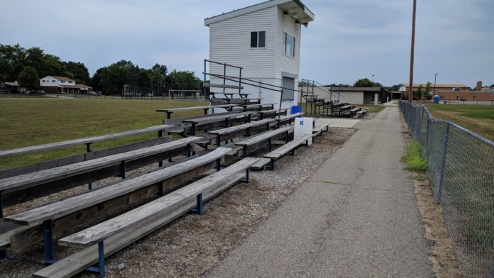 Outdoor athletic facility showing wooden bleacher seating with blue supports, press box structure, and sports field lighting