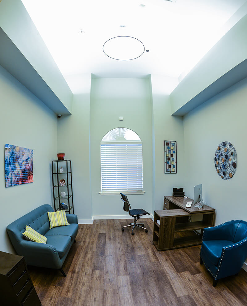 Interior professional office space with natural lighting, desk setup, comfortable seating, and therapeutic artwork on walls