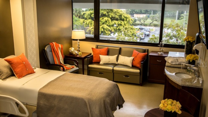 Interior patient bedroom with hospital bed, seating area with orange accents, window views, and en-suite bathroom with sink