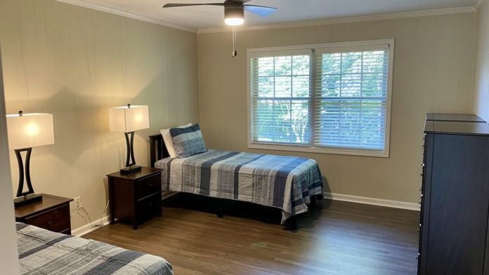 Interior patient bedroom with two single beds, patterned bedspreads, wooden nightstands with lamps, dresser, and large windows