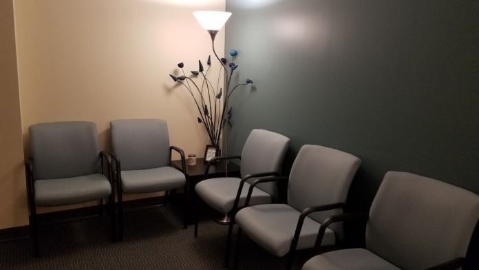 Interior waiting room with gray padded chairs, floor lamp, and decorative plant and artwork on walls