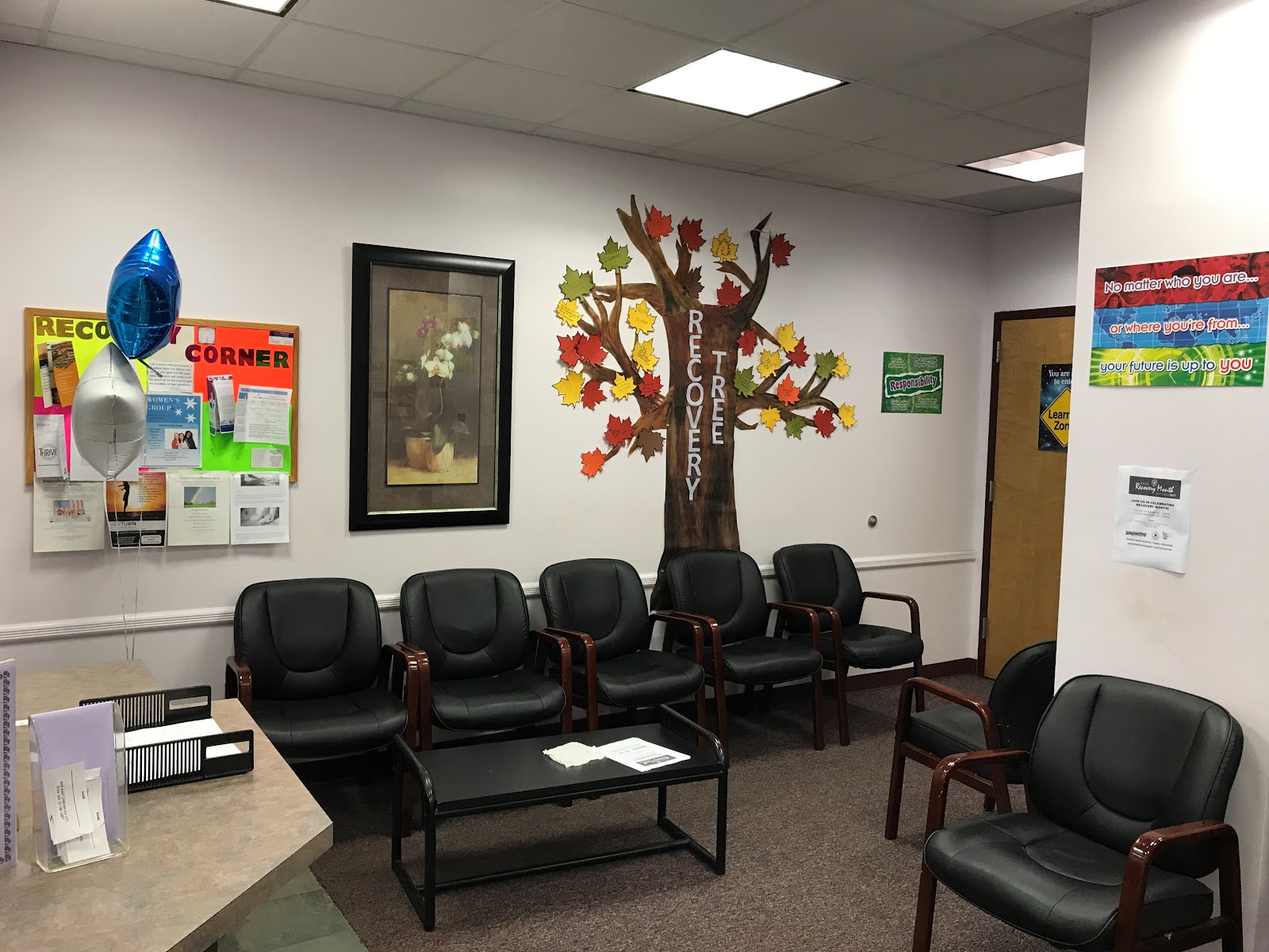 Interior waiting room with black leather chairs arranged in rows, white walls, and motivational tree decoration.