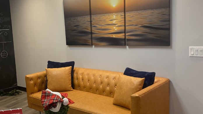Interior waiting room with gold tufted sofa, three-panel ocean sunset artwork above, white round table with decorative objects, and recessed ceiling lights