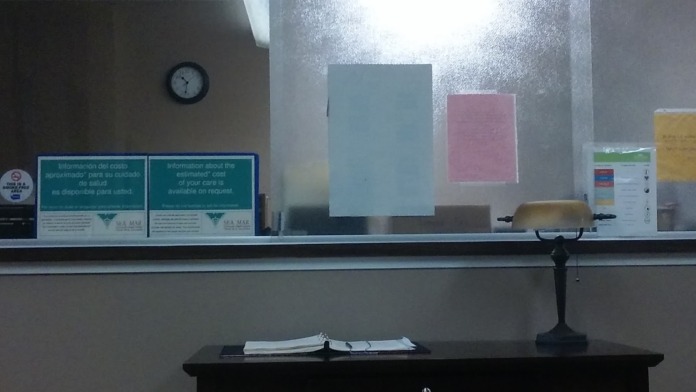 Interior office area showing reception window, clock on wall, and desk with materials and informational signage