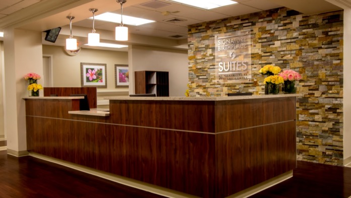 Interior reception area with dark wood curved desk, stone accent wall, pendant lighting, decorative flowers, and facility signage