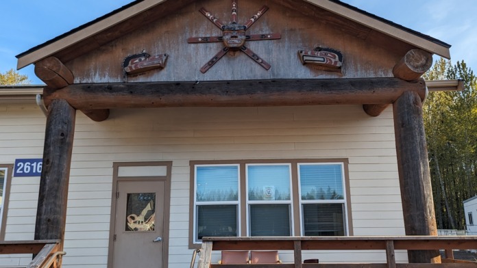 Exterior of single-story residential building with white siding, pitched roof with decorative sun carving, windows, and covered porch.
