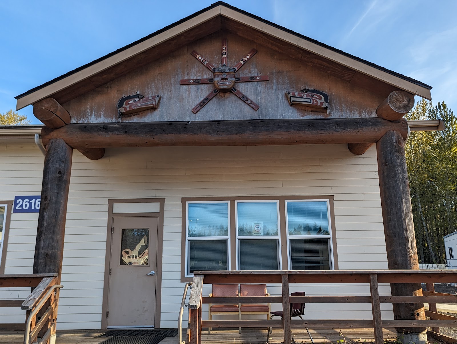 Exterior of single-story residential building with white siding, pitched roof with decorative sun carving, windows, and covered porch.