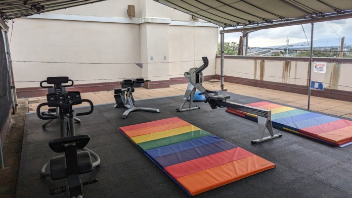 Outdoor covered rooftop area with exercise bikes, yoga mats, and colorful equipment with city and mountain views