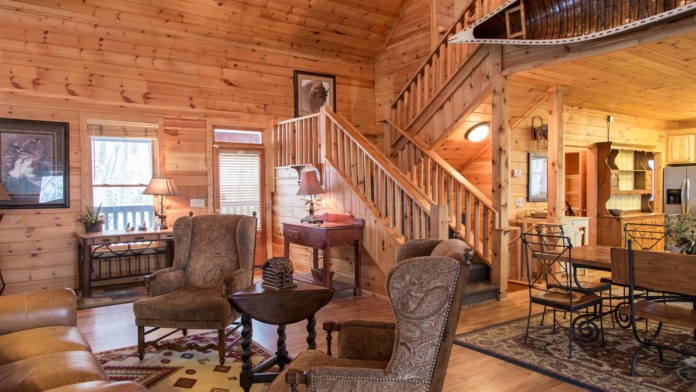 Interior of wooden lodge with vaulted cedar ceiling, fireplace, upholstered seating, staircase, and multiple windows providing natural light