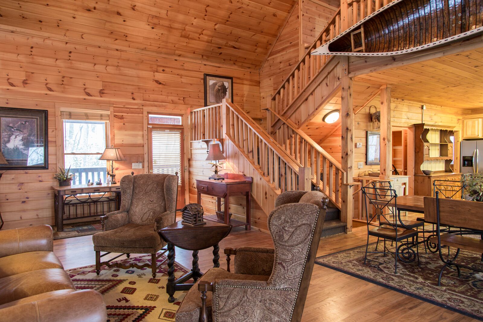 Interior of wooden lodge with vaulted cedar ceiling, fireplace, upholstered seating, staircase, and multiple windows providing natural light