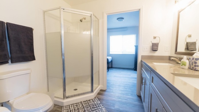Interior bathroom with walk-in shower, dual vanity, toilet, mirror, tile work, and doorway to bedroom