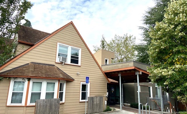Exterior view of facility building with tan and brown siding, entrance door, windows, and surrounding landscaping