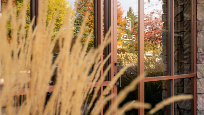 Exterior view of facility side entrance with green-framed glass doors, stone cladding, landscaping, and Zelus Recovery signage visible through glass