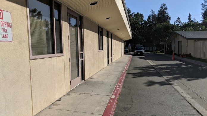 Side exterior view showing tan stucco facade, large windows, fire lane curb, parking area and surrounding landscape.