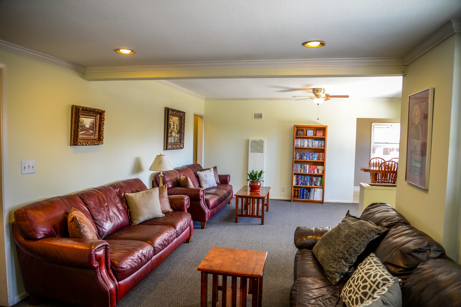 Interior common room featuring burgundy leather sofa, multiple comfortable seating areas, bookshelf, wall decorations, and natural window lighting