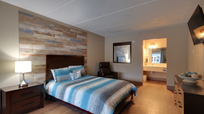 Interior patient bedroom with queen bed, striped bedding, wooden headboard, nightstand lamp, desk area, and bathroom vanity