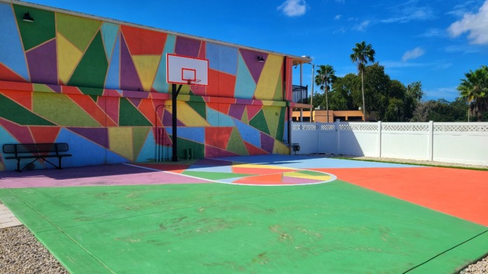 Exterior multi-sport court with vibrant geometric wall mural, basketball hoop, painted court surface, and palm trees