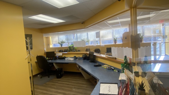 Interior office space with yellow walls, multiple desks, office chairs, and large windows with natural light