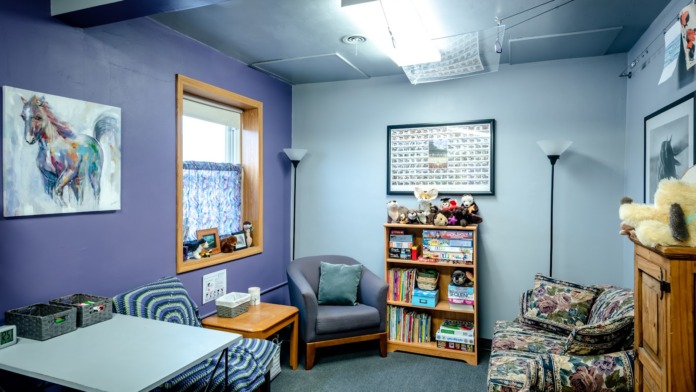 Interior therapy room with comfortable seating, wooden bookshelf with books and games, framed artwork, and purple and blue painted walls.