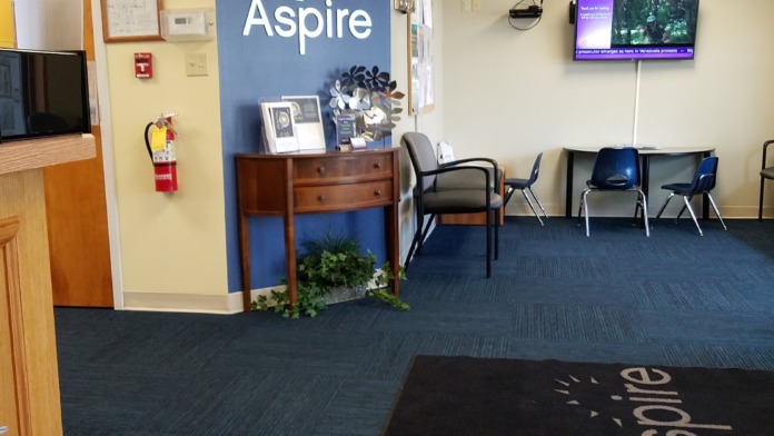 Interior waiting room with yellow walls, blue carpet, wooden reception desk, seating chairs, wall-mounted television, and Aspire branding