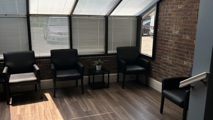 Interior seating area with glass walls, black vinyl chairs on wooden flooring, and exposed brick wall.