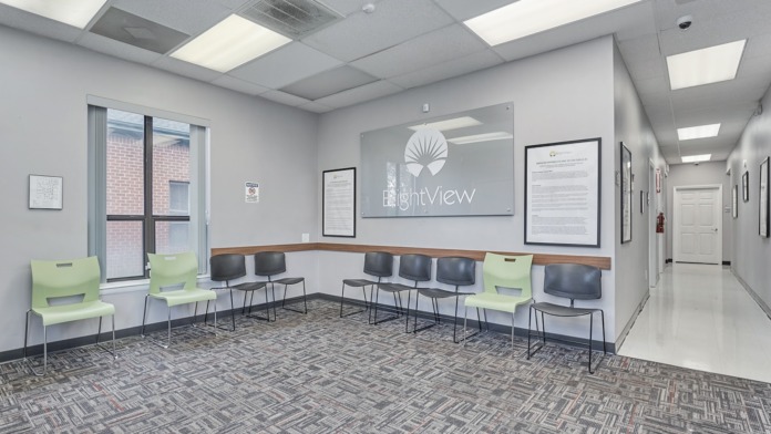 Interior waiting area with green and black chairs, ceiling lights, framed signage, and windows overlooking parking area
