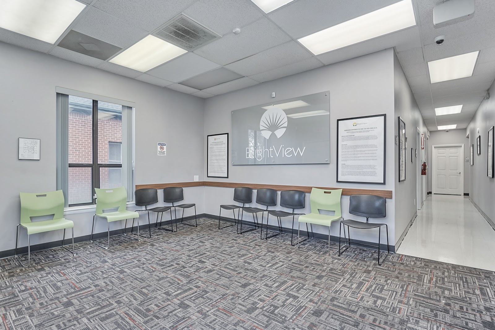 Interior waiting area with green and black chairs, ceiling lights, framed signage, and windows overlooking parking area