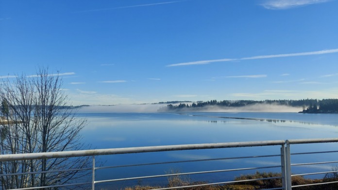 Outdoor scenic vista showing calm water, forested shoreline, and facility grounds with metal railing