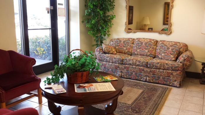 Interior reception area with upholstered seating, round wooden table with plant, mirror, and natural light from windows