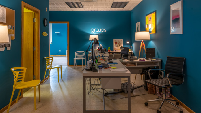 Interior office with teal walls, desk workstations, yellow and wood chairs, groups signage, yellow doors, framed artwork, modern furnishings