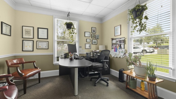 Interior office with round white table, office chair, leather seating, framed certifications, plants, and window view