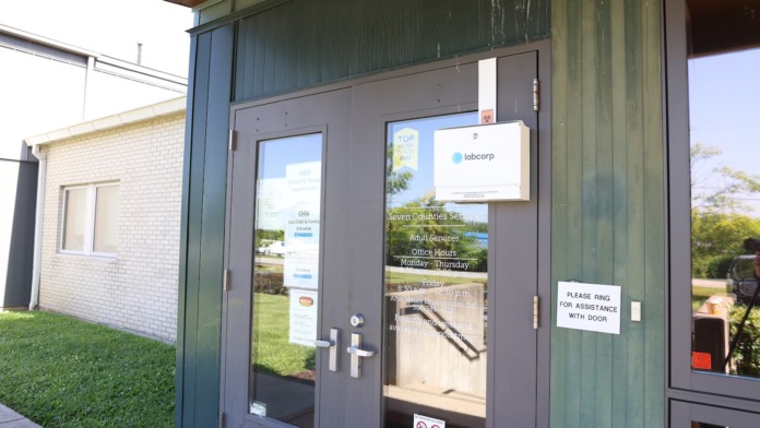 Exterior entrance of office building with glass doors, signage, and covered canopy near parking area and landscaping