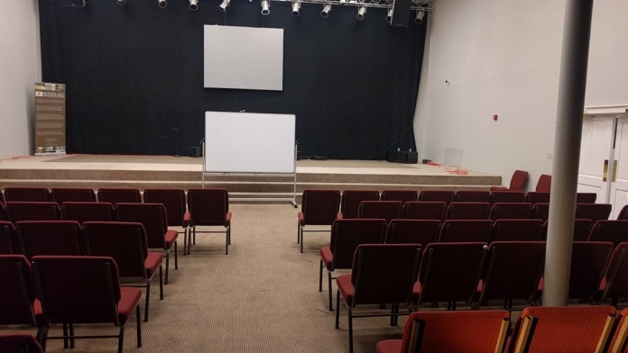 Interior auditorium with rows of burgundy theater seating, stage with projection screens, and support columns