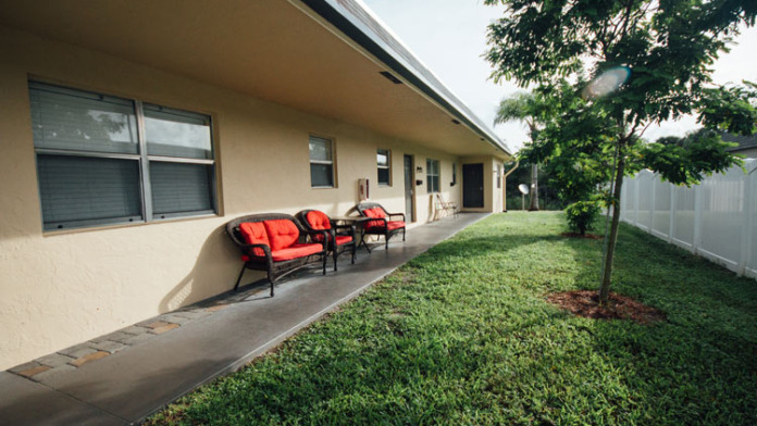 Exterior backyard area with red cushioned seating, shaded patio cover, green lawn, and white fence