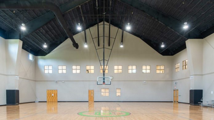 Interior gymnasium with wooden court, vaulted dark ceiling, basketball hoops, and glass windows