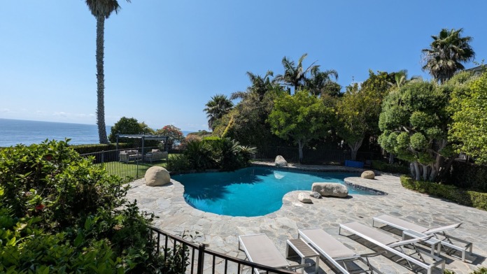 Exterior pool area with blue-water swimming pool, stone patio, loungers, and lush landscaping overlooking ocean