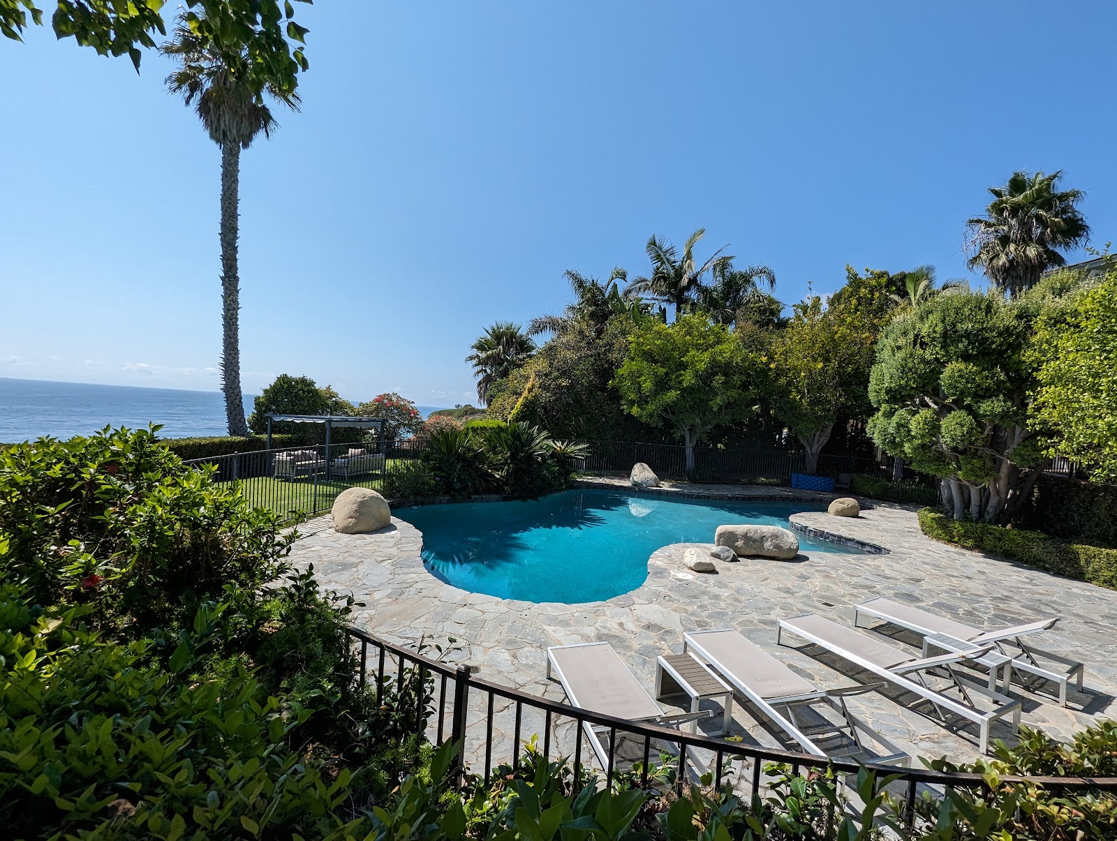 Exterior pool area with blue-water swimming pool, stone patio, loungers, and lush landscaping overlooking ocean