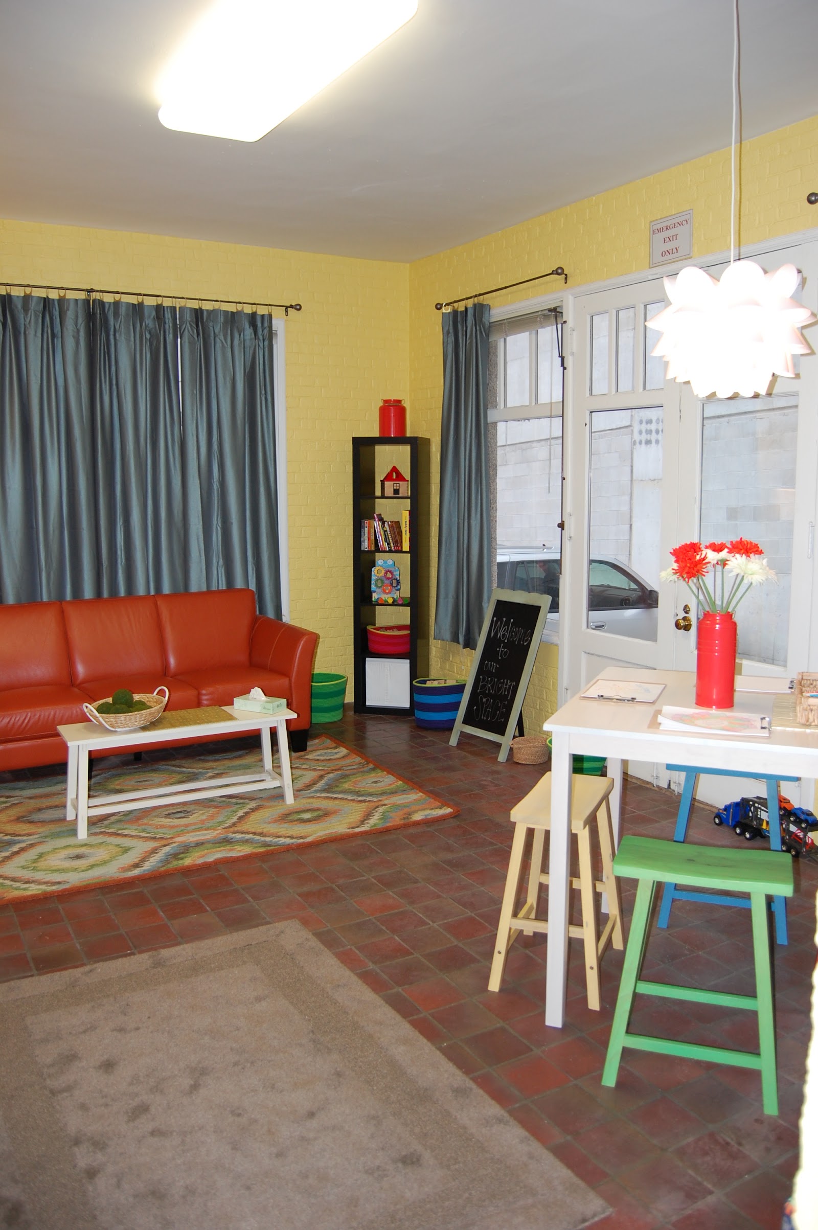 Interior therapy space with yellow walls, red seating, activity tables, shelving with toys, and colorful floor mats