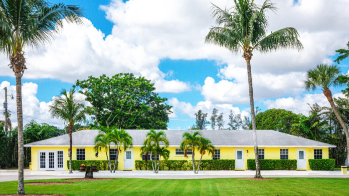 Exterior front view of bright yellow building with white roof, palm trees, manicured lawn, and clear blue sky