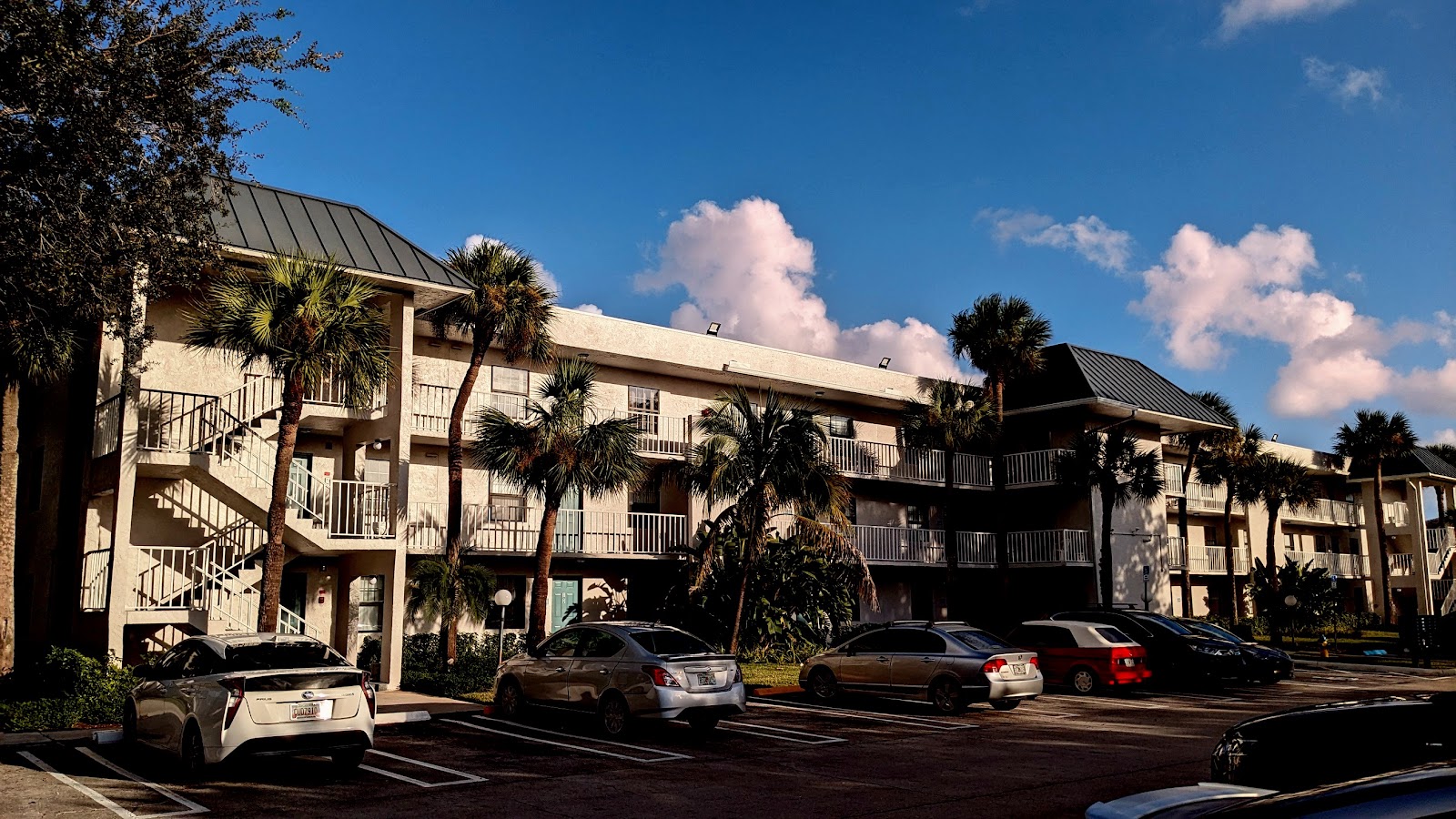 Exterior view of residential complex showing multi-unit buildings with metal railings, parking spaces, and surrounding trees