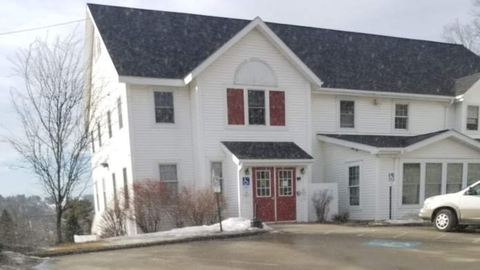 Exterior view of facility main entrance with red doors, white building facade, parking area with snow, and mature tree