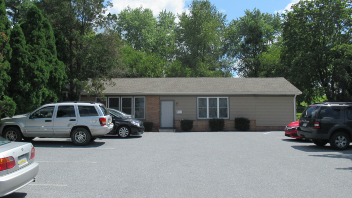 Exterior view of single-story facility building with brick base, siding, large windows, entrance door, and adjacent parking spaces with vehicles