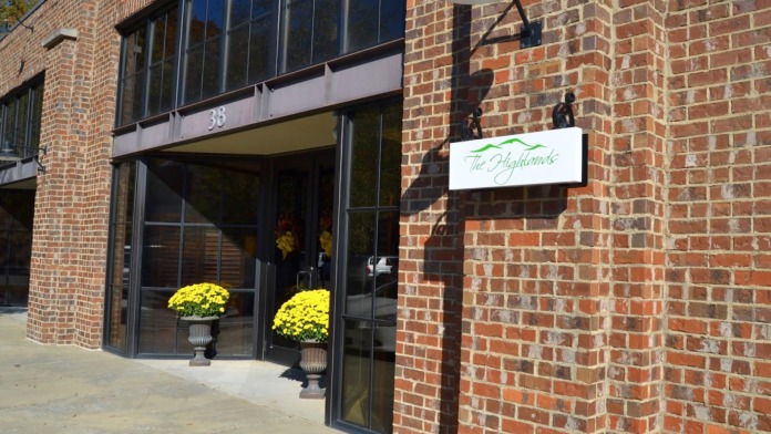 Exterior entrance showing brick building facade with The Highlands logo sign, large windows, and yellow flower planters