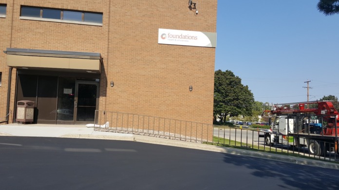 Exterior brick building facade with main entrance, signage, metal railings, and street-level parking area.