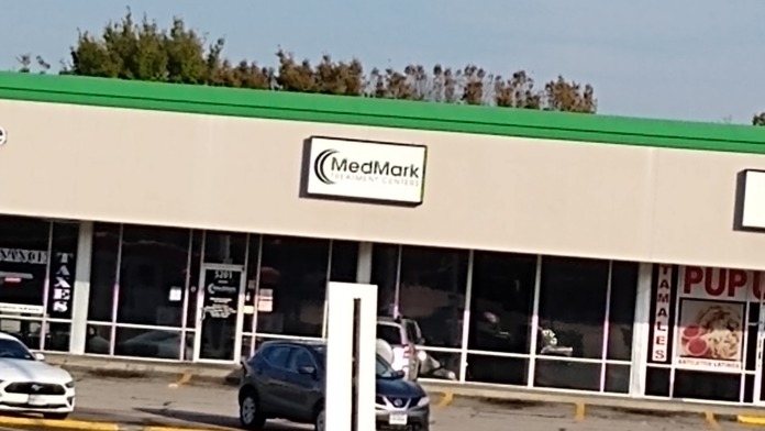 Exterior storefront of MedMark treatment center showing signage, large windows, green awning trim, and parking area with vehicles
