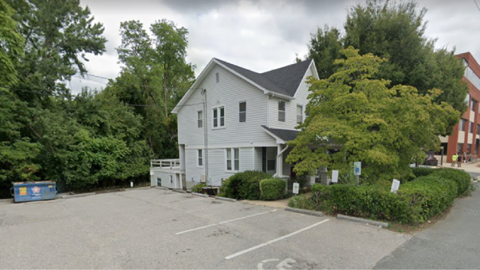 Exterior view of treatment facility showing white residential building with landscaping, trees, and adjacent brick building in background